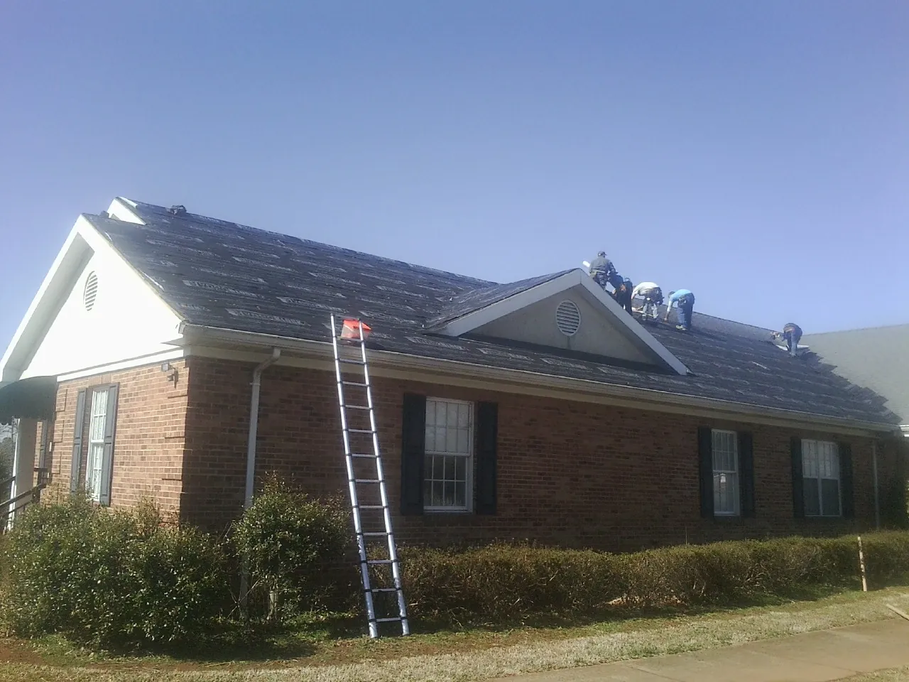 Crew tearing shingles off a brick ranch roof with a ladder set — Emergency Roof Repair work by our crew in Ames