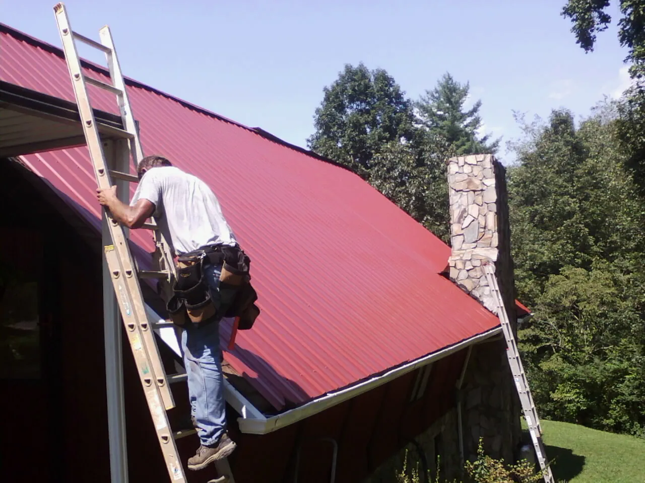 Roofer on a ladder beside a red metal roof and stone chimney — Soffit & Fascia Repair by Roofing Brothers in Ames